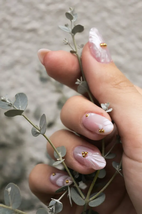 Unhas nude com aplicações douradas segurando um ramo de eucalipto