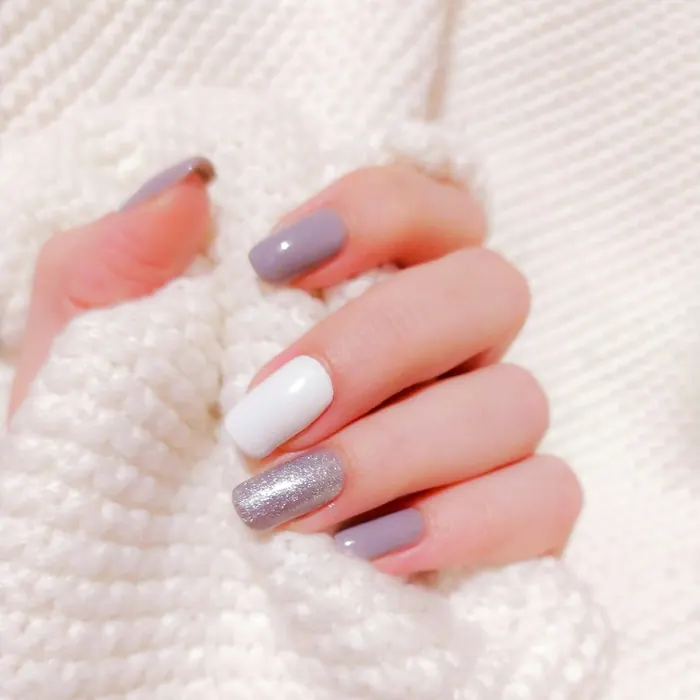 Mão com unhas lilás, brancas e com glitter prata segurando um suéter branco